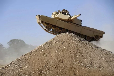 HD desktop wallpaper of an M1 Abrams military tank climbing a rocky hill, with a clear blue sky in the background.