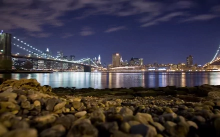 HD wallpaper of the Brooklyn Bridge and Manhattan skyline at night, with city lights reflecting on the water, captured from a rocky shoreline in New York.