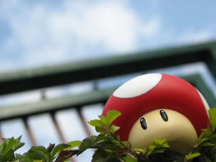 HD desktop wallpaper featuring a Super Mario Bros. mushroom toy peeking out from green foliage with a blurry background of a blue sky and a metal fence.