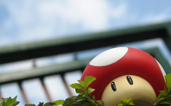 HD desktop wallpaper featuring a Super Mario Bros. mushroom toy peeking out from green foliage with a blurry background of a blue sky and a metal fence.