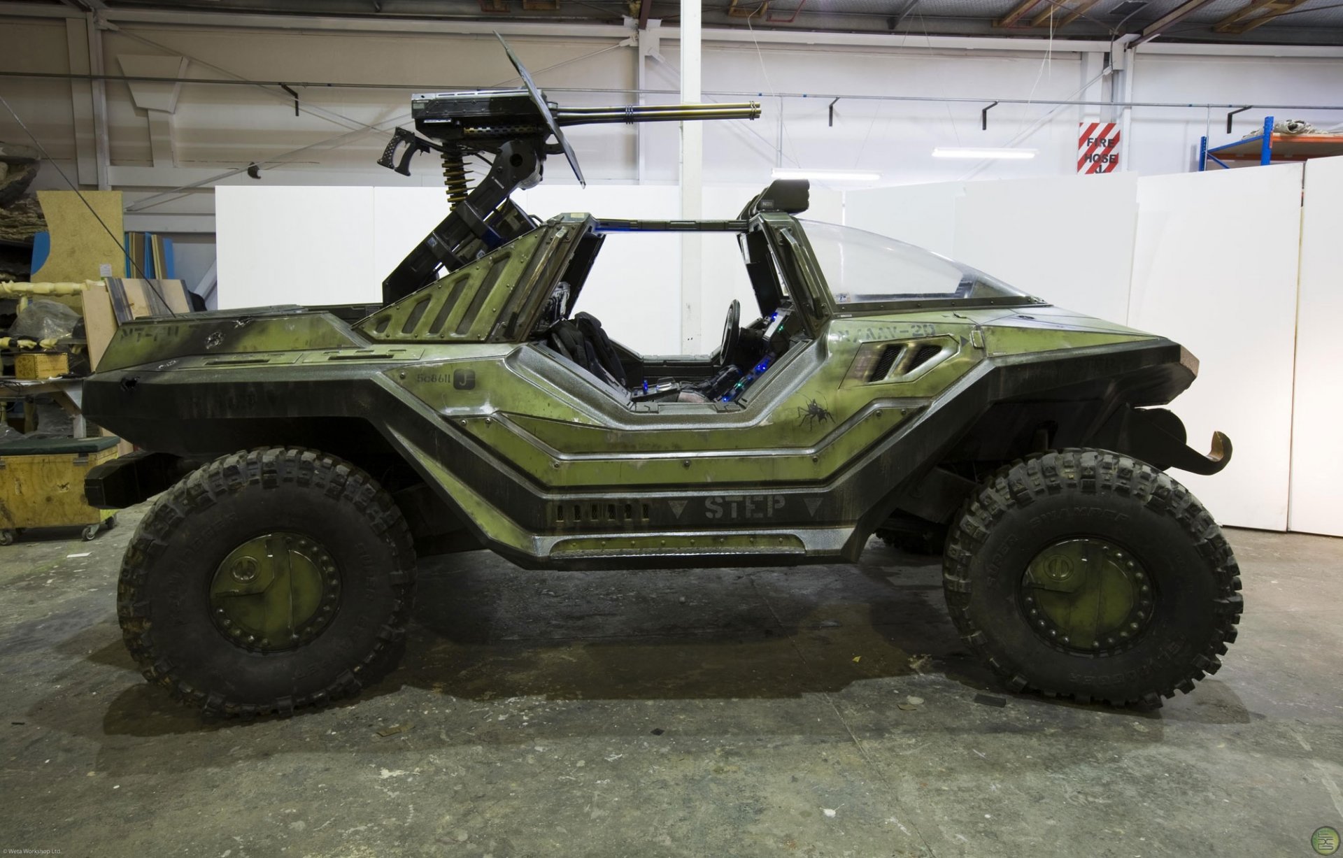 A high-definition desktop wallpaper featuring a military vehicle from the video game Halo, showcasing its rugged design and mounted weaponry in a garage environment.