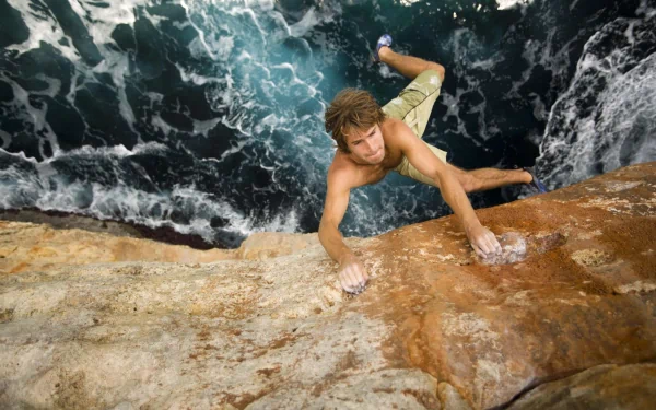 A climber ascends a rugged rock face above turbulent ocean waters, capturing the thrill of rock climbing in a dynamic HD desktop wallpaper.