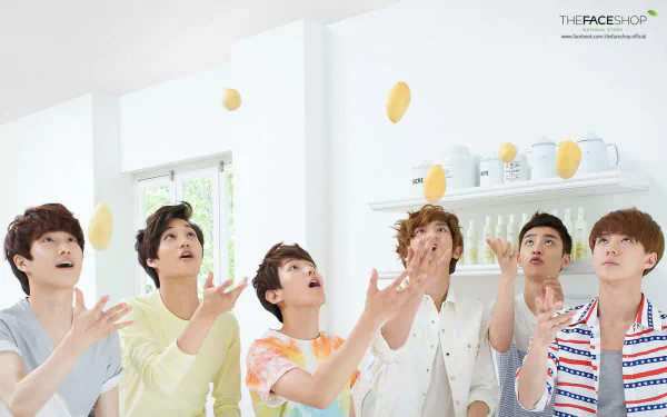 A vibrant HD desktop wallpaper featuring EXO members joyfully catching oranges in a bright, modern kitchen setting.