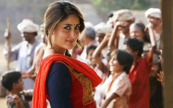 HD desktop wallpaper featuring Bollywood celebrity Kareena Kapoor. She is dressed in traditional attire with a red dupatta, smiling, and standing in front of a group of people in a rural setting.