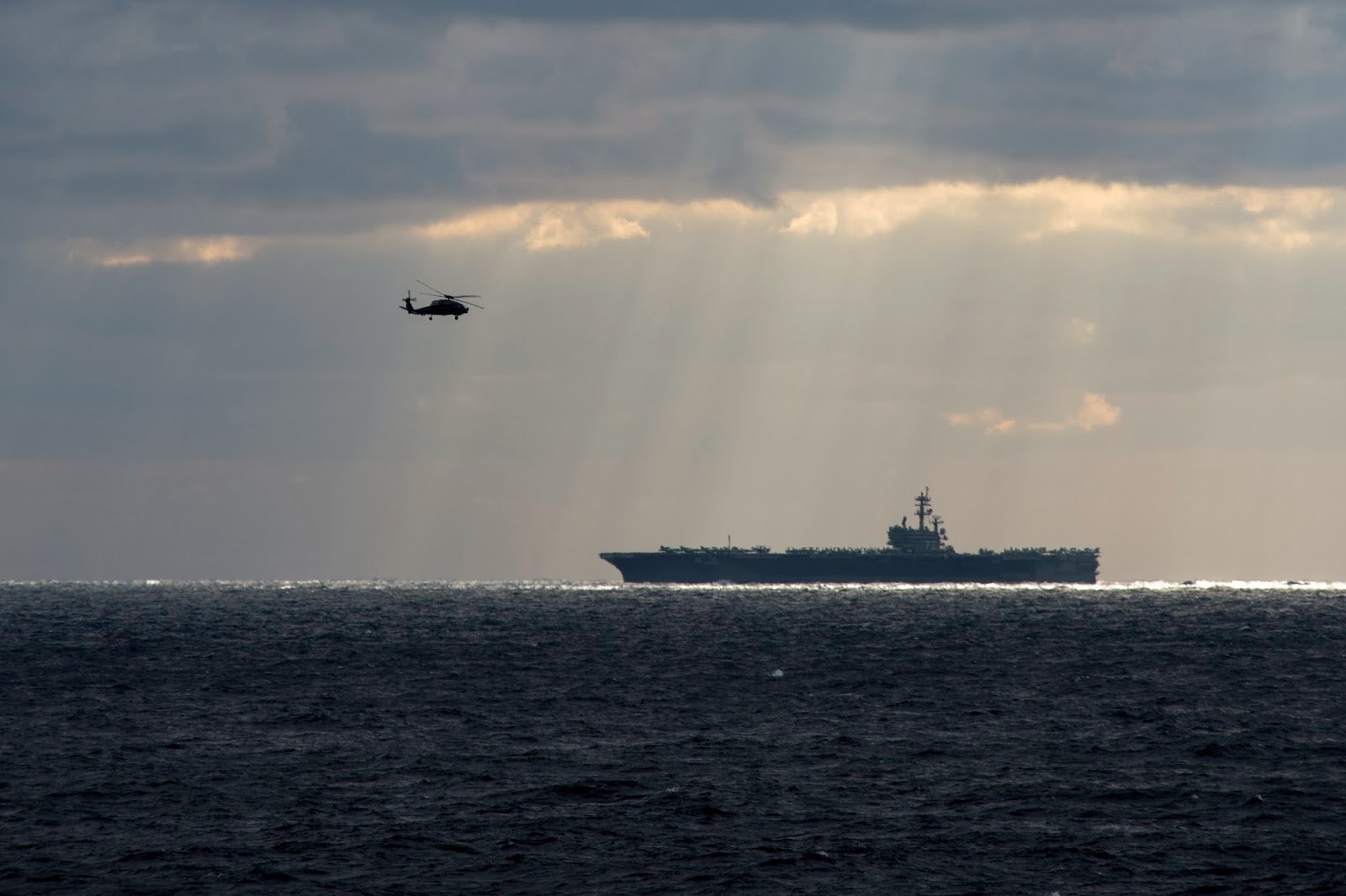 A helicopter hovers near the USS George H.W. Bush (CVN-77) aircraft carrier, illuminated by sunbeams, with calm waters under a dramatic sky.