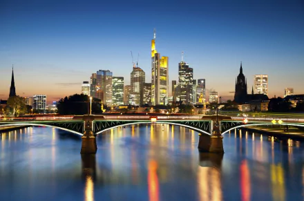 A 4K Ultra HD image of a man-made bridge in Frankfurt, Germany, with the illuminated city skyline reflected on the calm river at dusk.