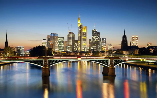 A 4K Ultra HD image of a man-made bridge in Frankfurt, Germany, with the illuminated city skyline reflected on the calm river at dusk.