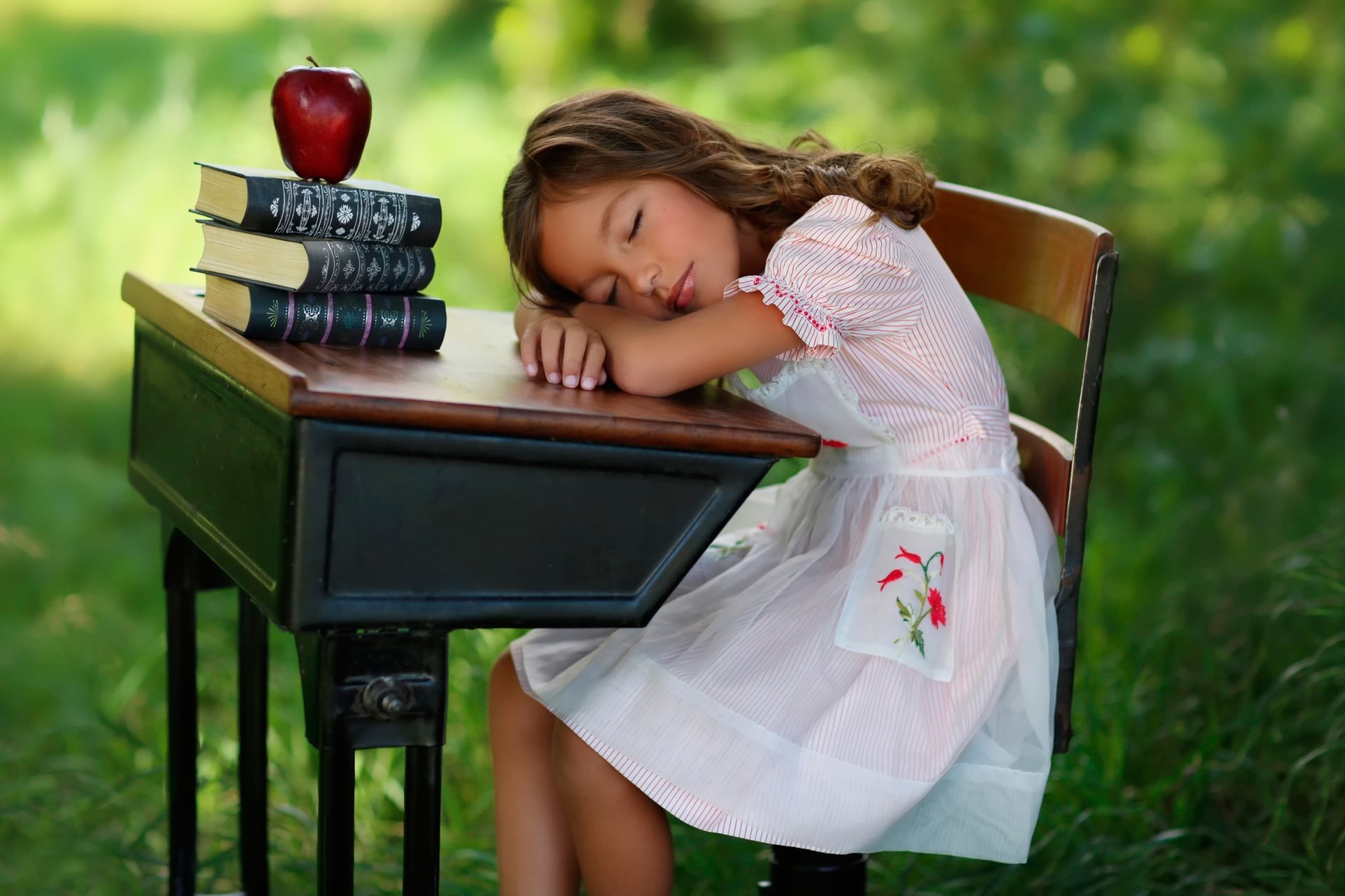 A little girl rests her head on a vintage wooden desk with stacked books and an apple, surrounded by lush greenery. This HD summer-themed wallpaper offers a serene and nostalgic moment of childhood.