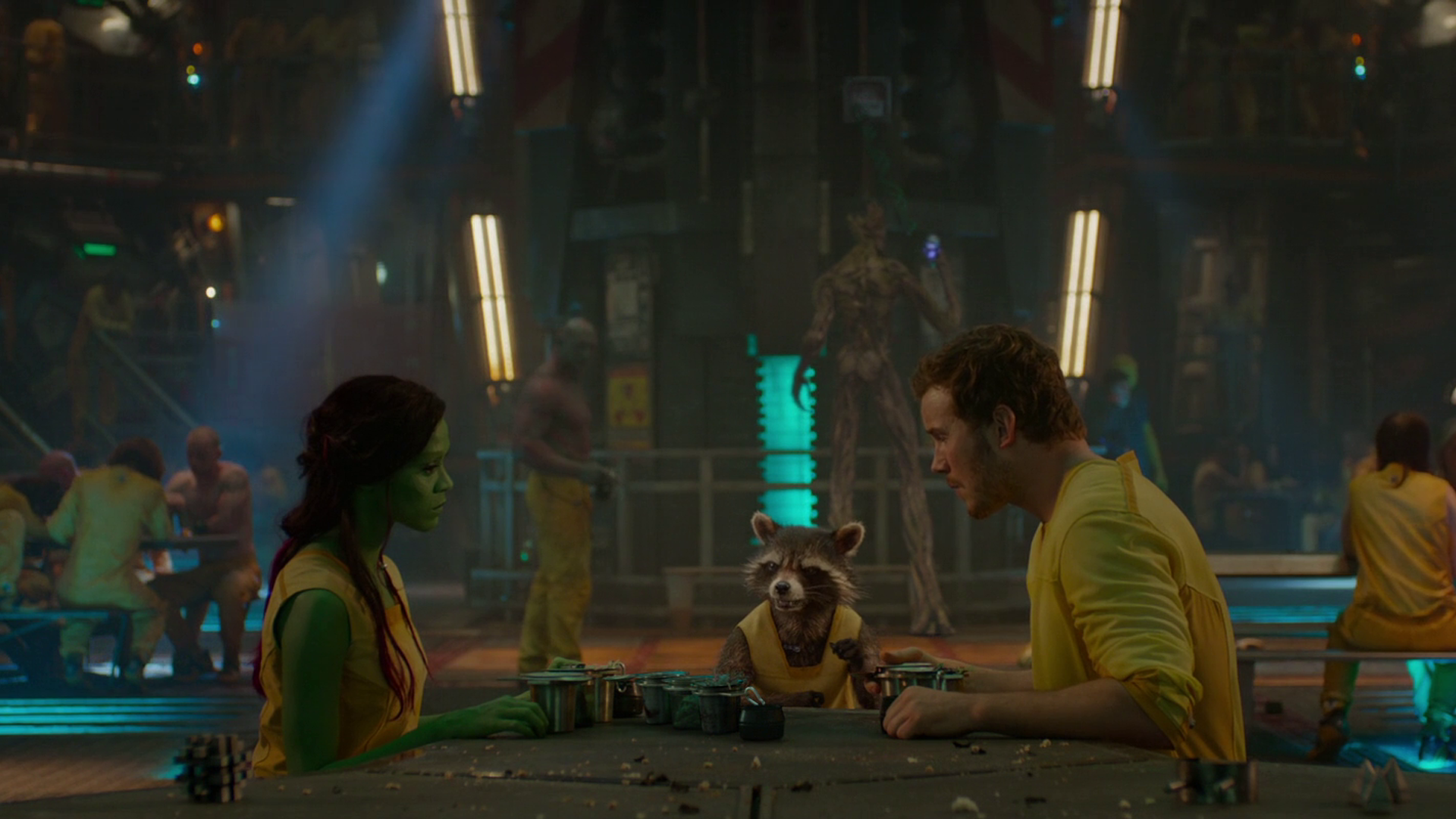 Zoe Saldana and Chris Pratt as Gamora and Peter Quill sit across from Rocket Raccoon in a vibrant scene from *Guardians of the Galaxy*, featuring lively background characters.