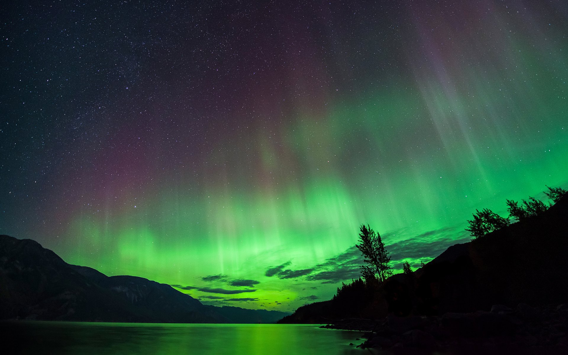 HD desktop wallpaper capturing a night scene of vibrant aurora borealis above a tranquil lake surrounded by silhouetted trees and mountains.