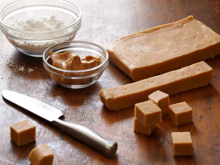 Close-up of peanut butter fudge squares with a knife, a bowl of peanut butter, and a bowl of powdered sugar on a wooden surface, rendered in 4K Ultra HD for desktop wallpaper.