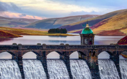 A stunning HD wallpaper showcasing a man-made dam with cascading water, framed by rolling hills and a serene landscape under a dramatic sky.