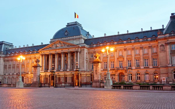 HD desktop wallpaper showcasing the Royal Palace of Brussels at dusk, illuminated under soft evening light with the Belgian flag atop the grand man-made historic building.
