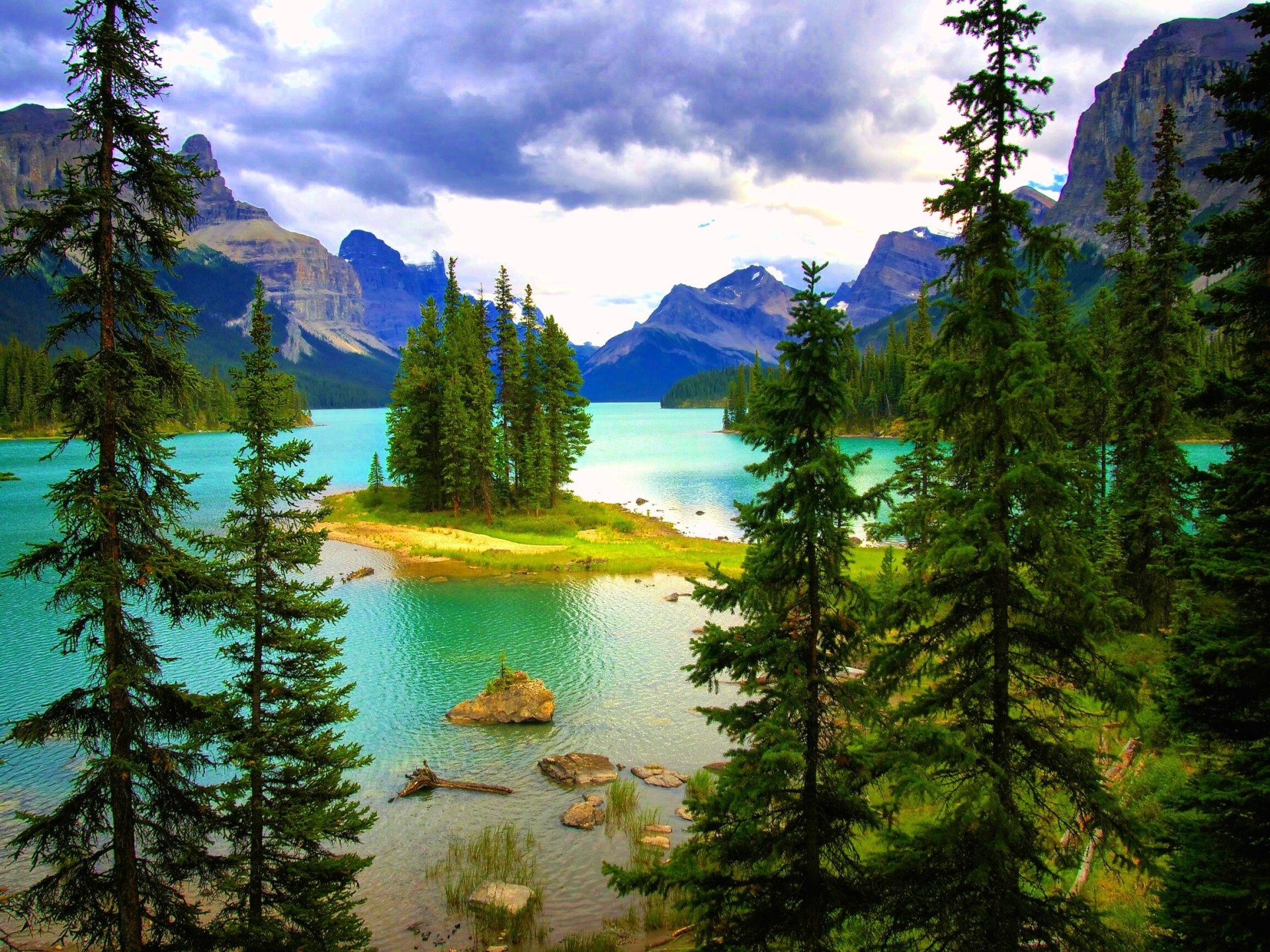 HD desktop wallpaper of a serene lake with a pine-covered islet, surrounded by towering mountains and lush nature under a partly cloudy sky.