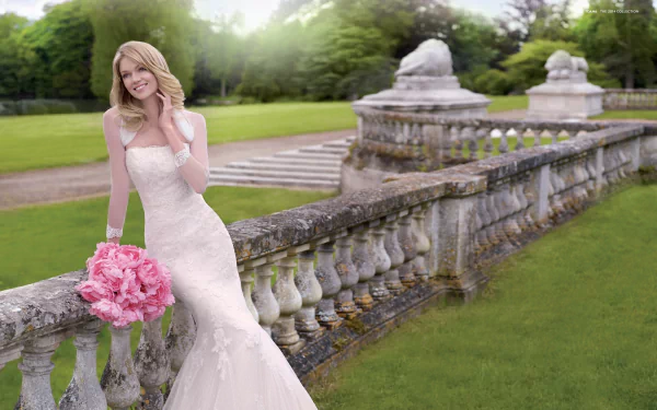 A woman in a stunning wedding dress holds a bouquet of pink flowers, posing gracefully near a stone railing in a lush garden, surrounded by greenery.