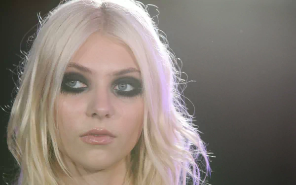 Close-up portrait of a blonde rock musician with heavy smoky eye makeup and pale lips, lit from behind — 2K Quad HD PC desktop wallpaper.