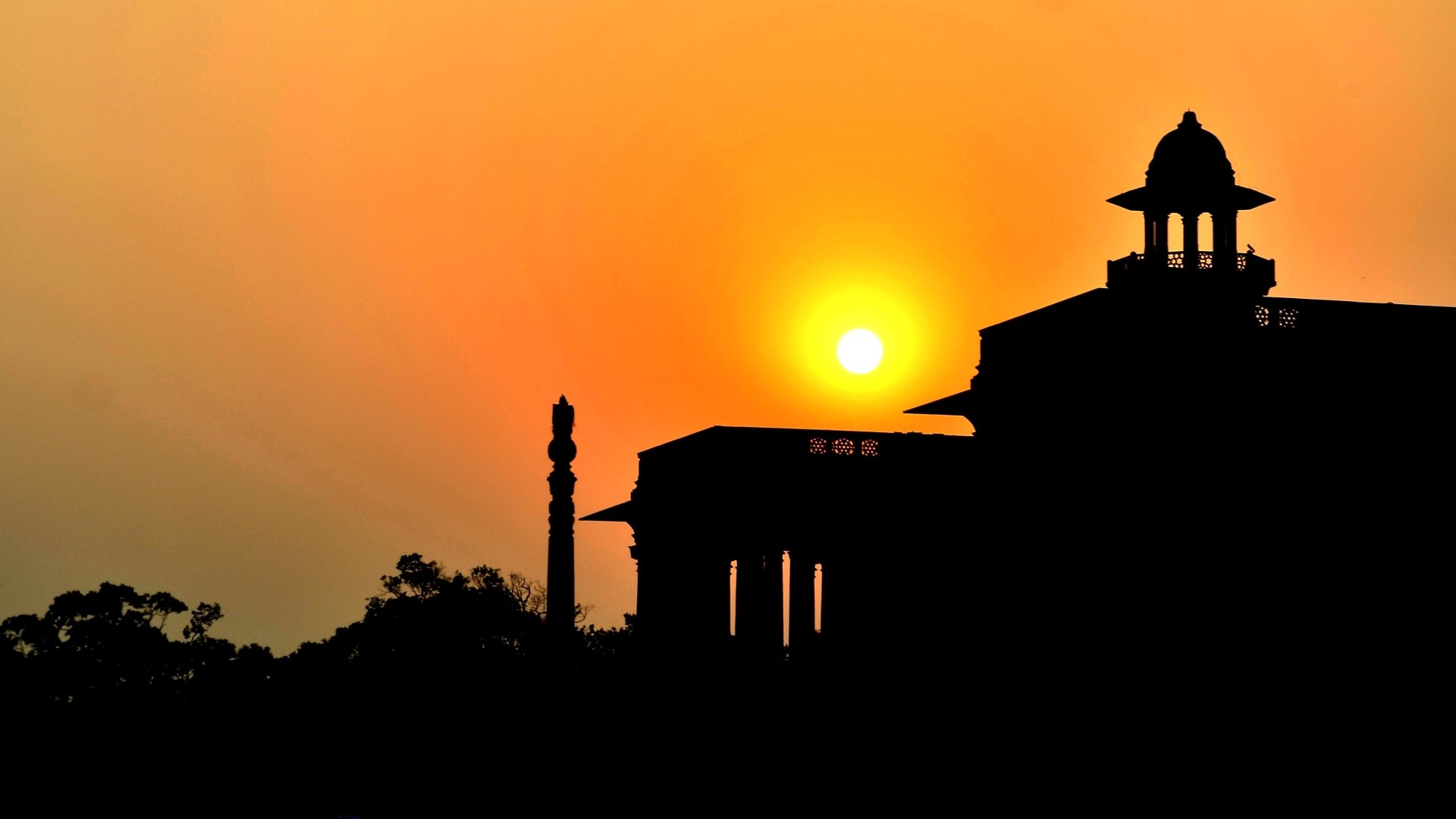 Silhouetted Rashtrapati Bhavan at sunset with a glowing sun and orange sky — man-made architecture rendered as an HD PC desktop wallpaper background.