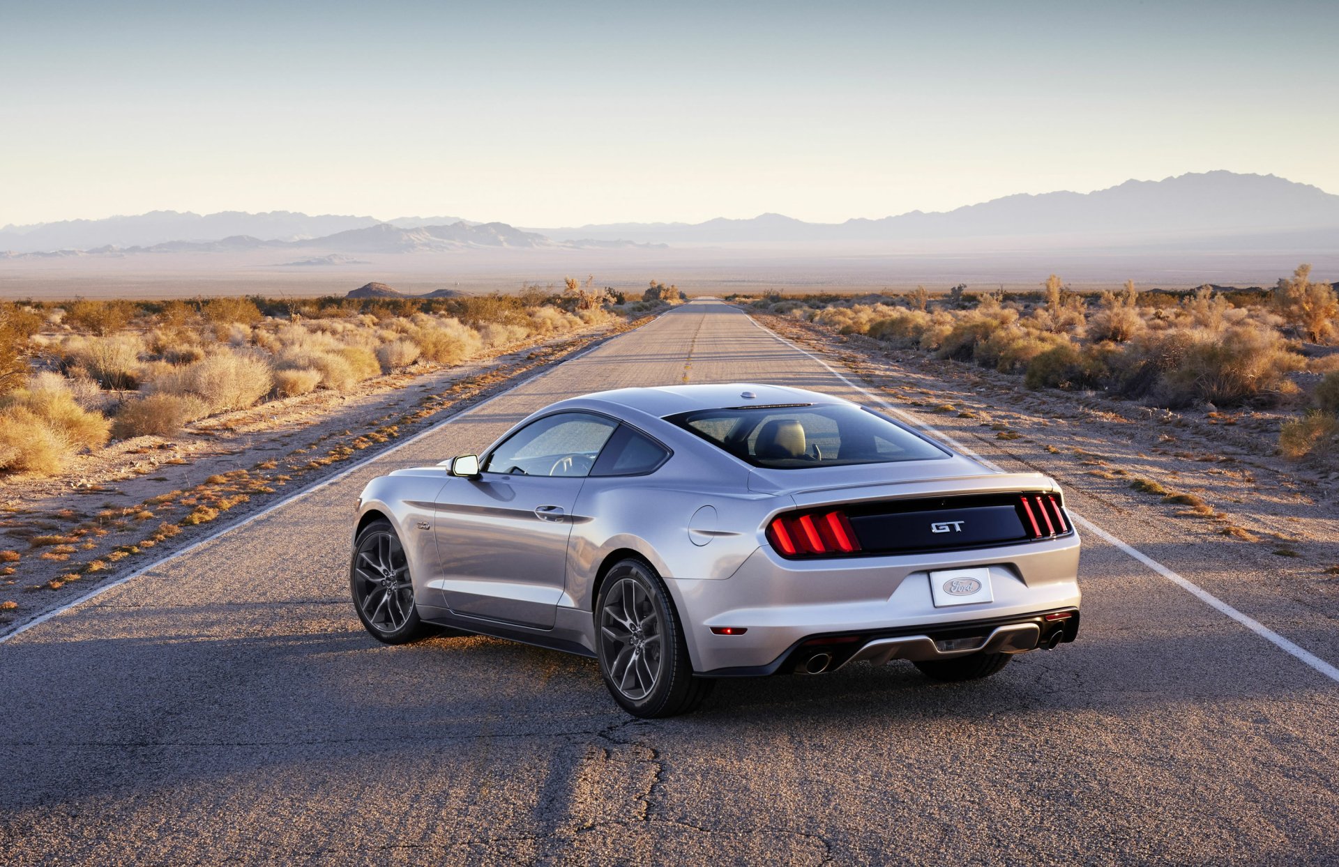 A sleek silver 2015 Ford Mustang GT parked on an empty desert road, with mountains in the background, showcasing a stunning HD wallpaper view.