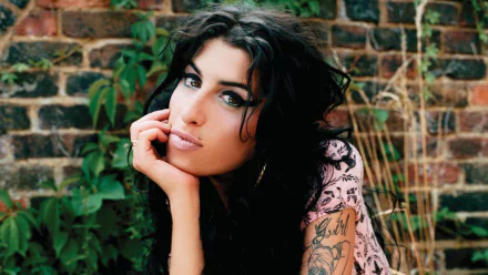 HD desktop wallpaper of English singer Amy Winehouse with dark hair and tattoos, posing thoughtfully against a brick wall with greenery.