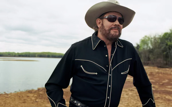 4K Ultra HD PC desktop wallpaper: country musician in a cowboy hat and sunglasses, wearing a black western shirt, standing by a lakeshore.