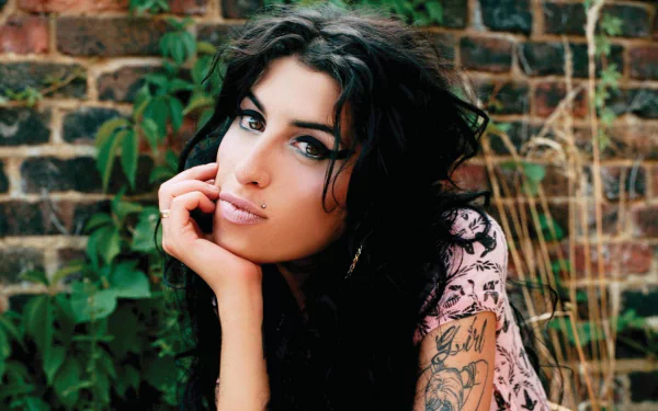 HD desktop wallpaper of English singer Amy Winehouse with dark hair and tattoos, posing thoughtfully against a brick wall with greenery.