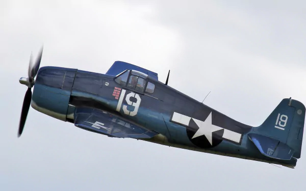 HD PC desktop wallpaper of a military Grumman F6F Hellcat in blue WWII markings banking across a cloudy sky
