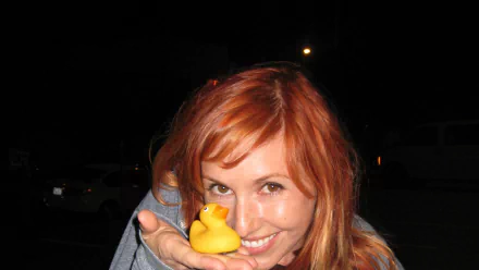 Close-up of a smiling red-haired woman holding a yellow rubber duck at night; 2K Quad HD PC desktop wallpaper/background.