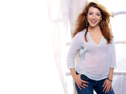 Alyson Hannigan poses with a warm smile, dressed in a light sweater and jeans, set against a bright, airy background. This HD desktop wallpaper captures her vibrant personality.