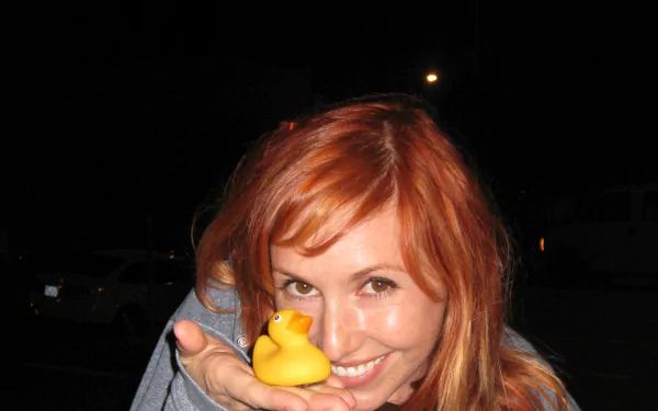 Close-up of a smiling red-haired woman holding a yellow rubber duck at night; 2K Quad HD PC desktop wallpaper/background.