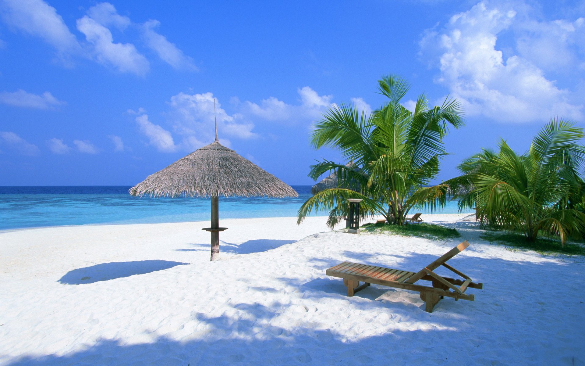 HD desktop wallpaper featuring a serene beach scene with white sand, clear blue sky, a thatched umbrella, palm trees, and wooden lounge chairs by the ocean.