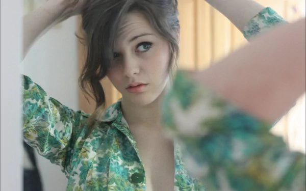 A brunette woman adjusts her hair in front of a mirror, dressed in a green floral shirt. The soft lighting creates a gentle reflection, enhancing the serene atmosphere.