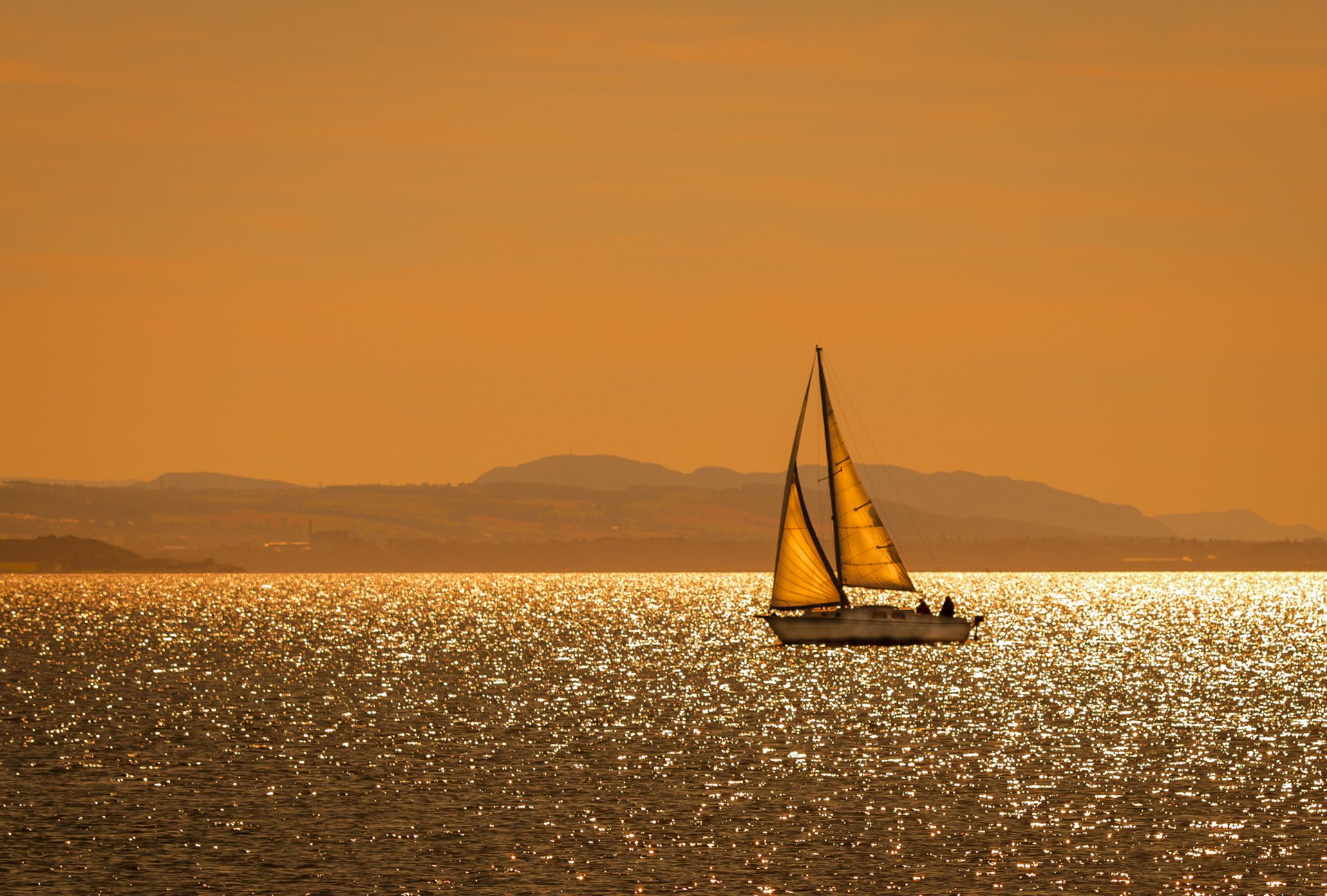 Sailboat boat/vehicle glides across a glittering orange sea at sunset, distant hills on the horizon — HD PC desktop wallpaper and background.