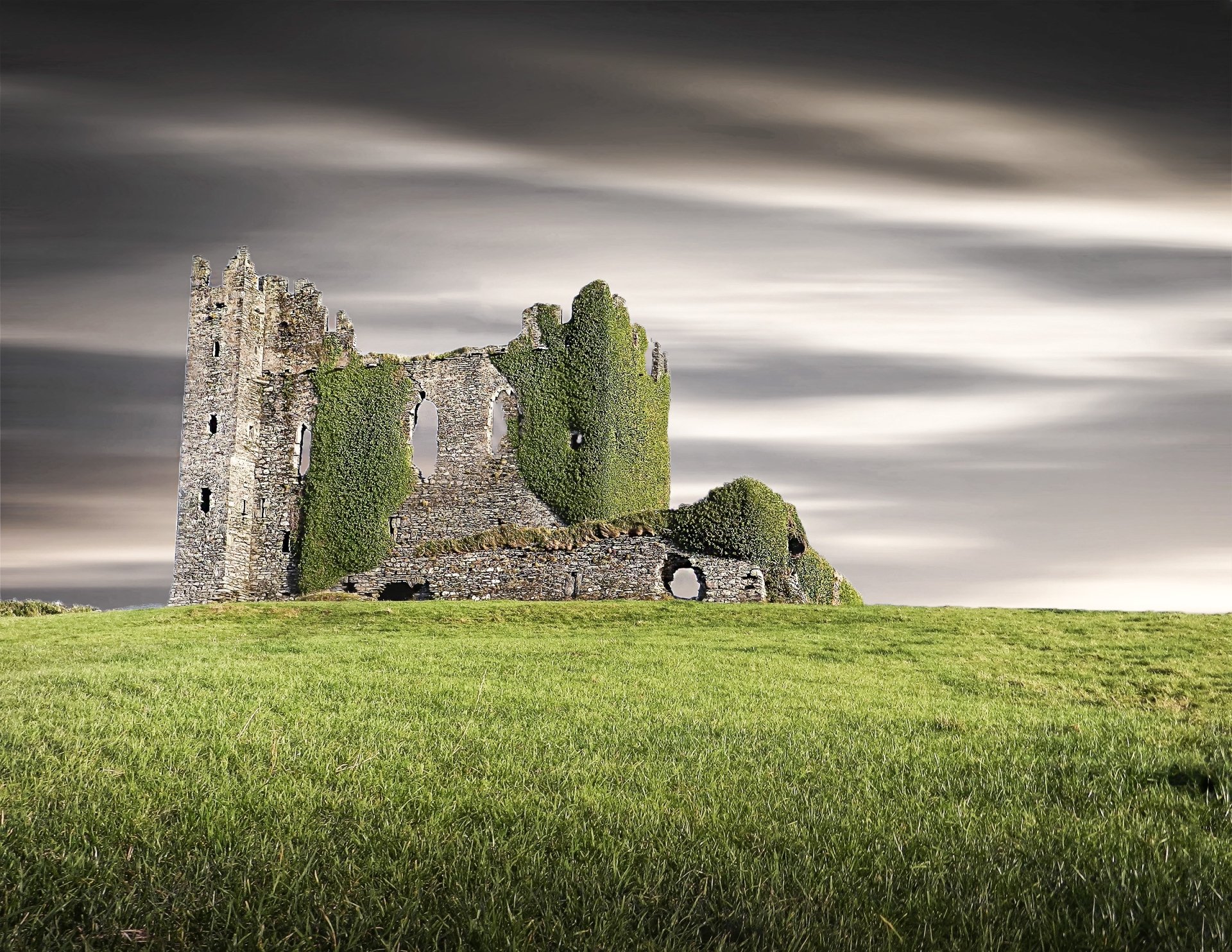 Ballycarbery Castle ruins, a man-made ivy-covered stone keep on a green field beneath dramatic clouds — HD PC desktop wallpaper/background.