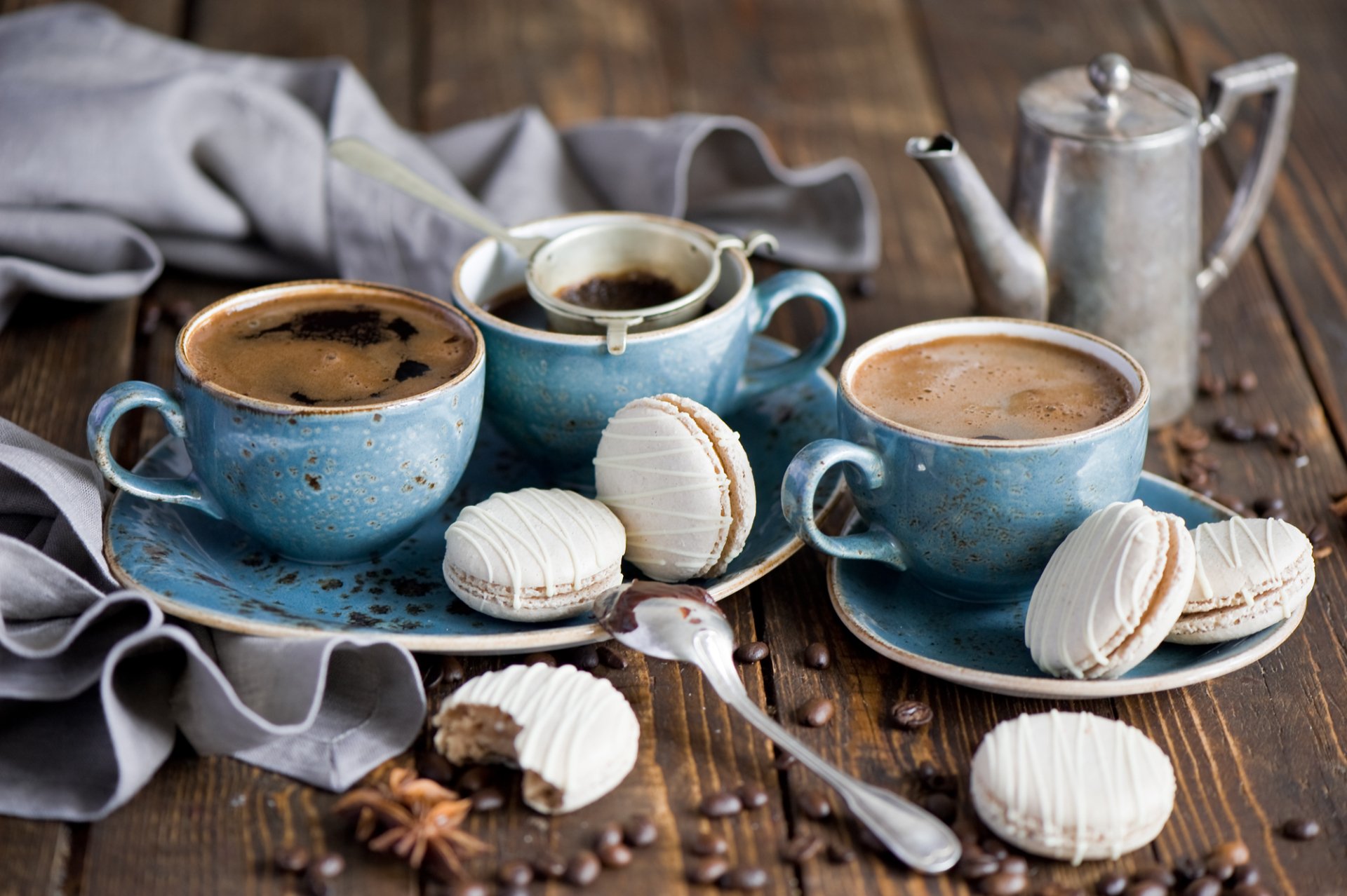 HD PC desktop wallpaper featuring three blue cups of coffee with white macarons on a rustic wooden table, surrounded by a silver coffee pot and a gray cloth.