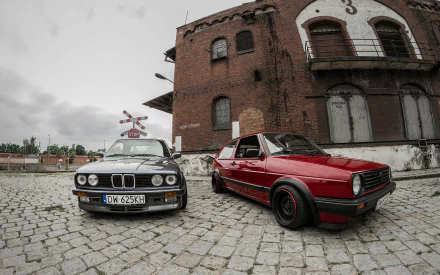 HD PC desktop wallpaper featuring a red Volkswagen Golf and a black BMW parked on a cobblestone street in front of a rustic brick building.