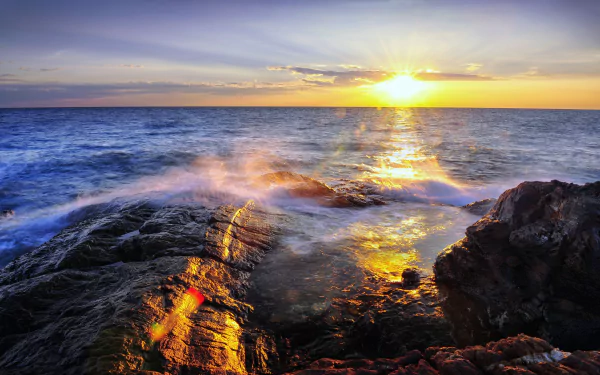 Vibrant 4K Ultra HD seaside sunset with the sun dipping below the horizon, casting golden light over rocky seashore and shimmering sea under a serene sky.
