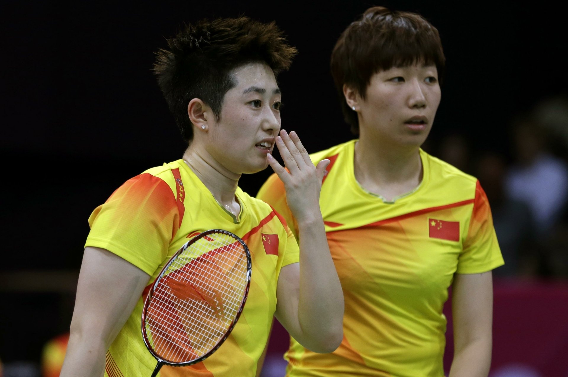 Two badminton players in yellow team uniforms discuss strategy. The image is an HD desktop wallpaper and background that showcases the sport of badminton.