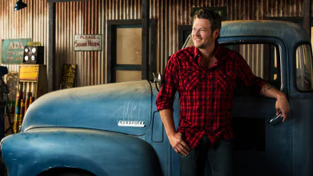 2K Quad HD PC desktop wallpaper of a smiling country singer in a red plaid shirt leaning on a vintage Chevrolet pickup inside a rustic music-themed garage.