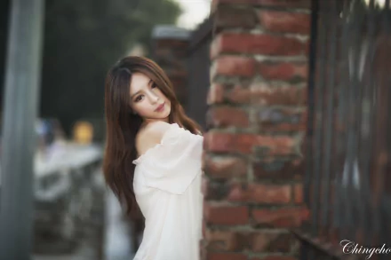 A woman in a white off-shoulder dress leans against a brick fence with a soft bokeh background, captured in a 4K Ultra HD image by Julie Chang and Zhang Qi Jun.