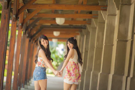 Two Asian women holding hands and smiling, standing under a corridor with wooden beams. The image is an HD desktop wallpaper and background.