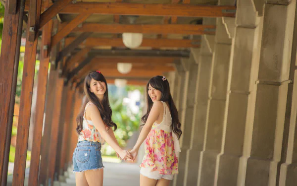 Two Asian women holding hands and smiling, standing under a corridor with wooden beams. The image is an HD desktop wallpaper and background.