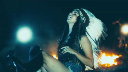Lana Del Rey smiles joyfully in a headdress, surrounded by a warm glow from a fire, capturing a vibrant and artistic moment in this HD music-themed desktop wallpaper.