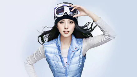A fashionable HD wallpaper featuring Celebrity Fan Bingbing with long hair, dressed in a stylish blue vest and winter accessories, set against a soft pastel background.