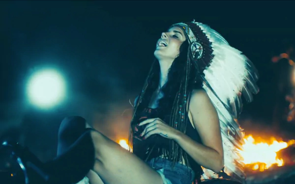 Lana Del Rey smiles joyfully in a headdress, surrounded by a warm glow from a fire, capturing a vibrant and artistic moment in this HD music-themed desktop wallpaper.