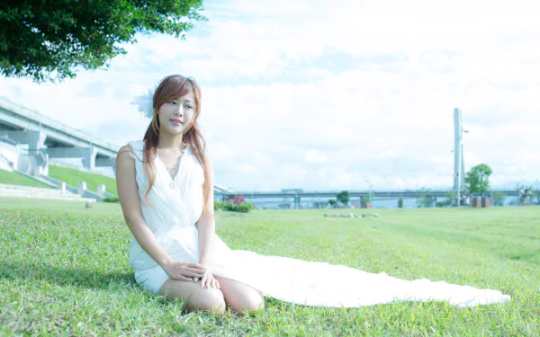A Taiwanese woman, Xiao Xi, in a white dress sits on a grassy field, with a scenic background of a bridge and trees. The image is a high-definition wallpaper and background.