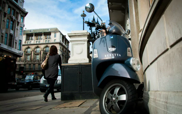 Blue Vespa scooter parked on a London, England, United Kingdom sidewalk amid man-made historic buildings; pedestrian passes by — HD PC desktop wallpaper background.
