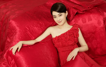 Smiling woman in a red silk one-shoulder dress reclines on a red sofa with matching pillows; vivid 5K Ultra HD PC desktop wallpaper and background.