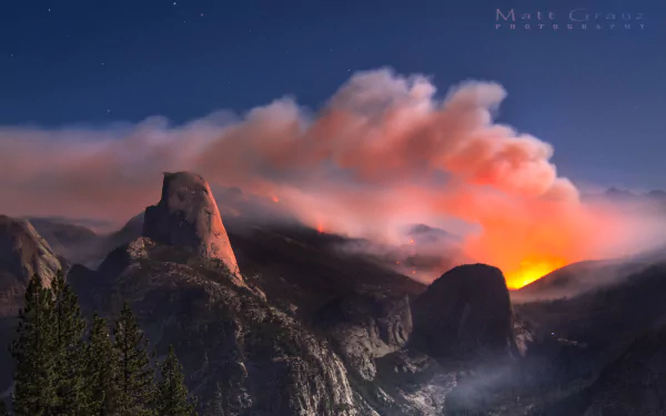 Nighttime view of Half Dome with glowing fire and smoke rising above the rugged landscape in this HD photography wallpaper.