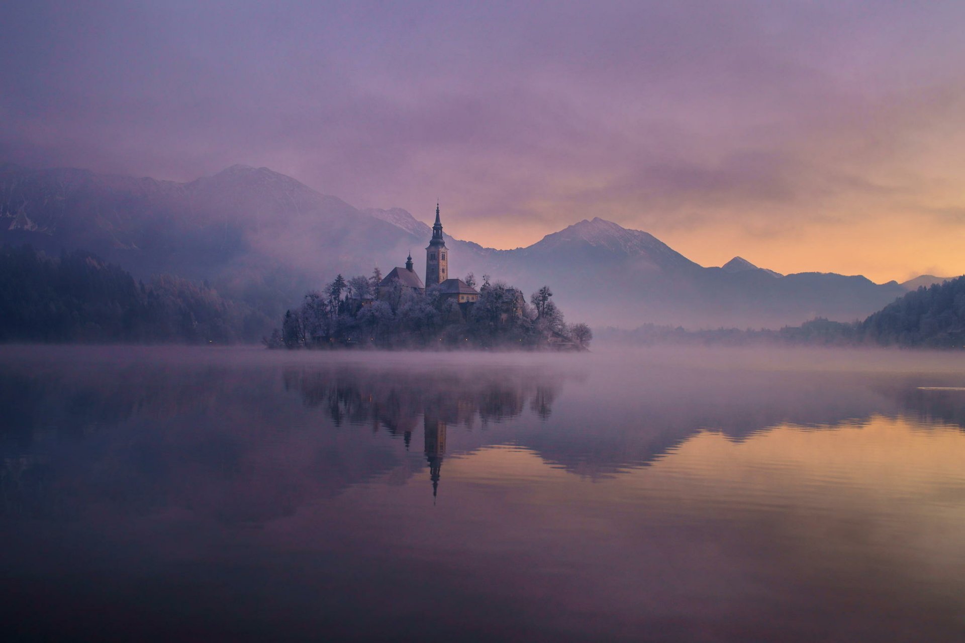 HD wallpaper of the Assumption of Mary Church on a misty island, surrounded by calm water and mountains under a colorful twilight sky.