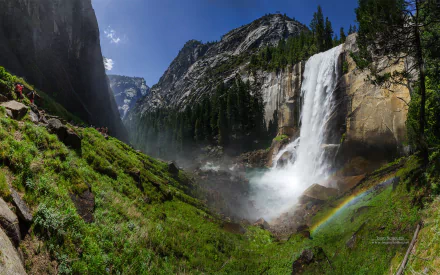 Vernal Fall cascades into the Merced River amid lush trees and towering mountains in Yosemite National Park, California, with a vibrant rainbow arching through the mist.